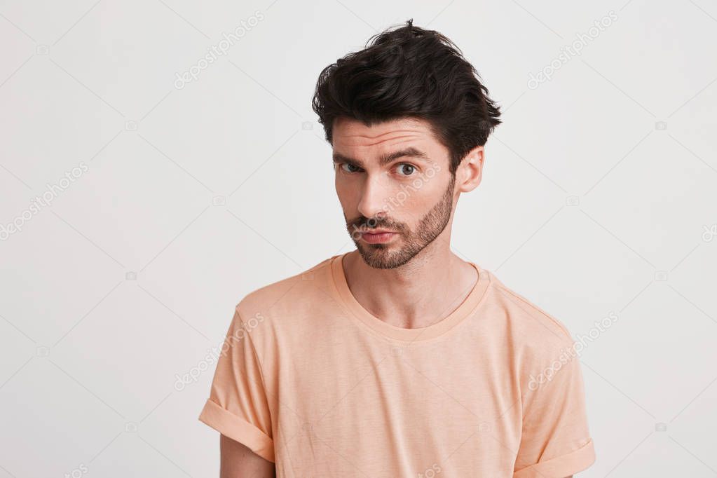 Closeup of serious handsome young man with bristle wears peach t shirt feels confident and looks directly in camera isolated over white background