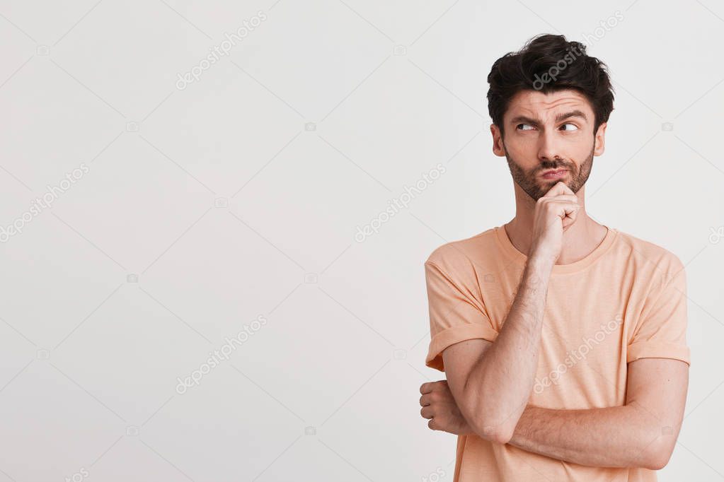 Portrait of serious thoughtful young man with bristle wears peach t shirt looks pensive and keeps hands folded isolated over white background