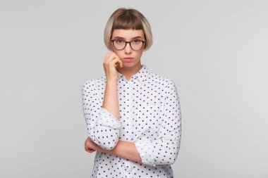 Düşünceli güzel sarışın genç kadın portresi puantiyeli gömlek giyer ve gözlük görünüyor konsantre ve tutar eller izole beyaz arka plan üzerinde katlanmış