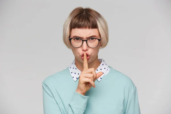 Closeup of serious strict young woman wears blue sweatshirt and spectacles looks displeased and shows silence gesture isolated over white background