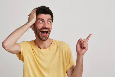 Portresi kıl ile neşeli genç adam hayret ve sarı tişörtlü heyecanlı görünüyor ve beyaz arka plan üzerinde izole parmak ile tarafına puan ağız giyer açıldı