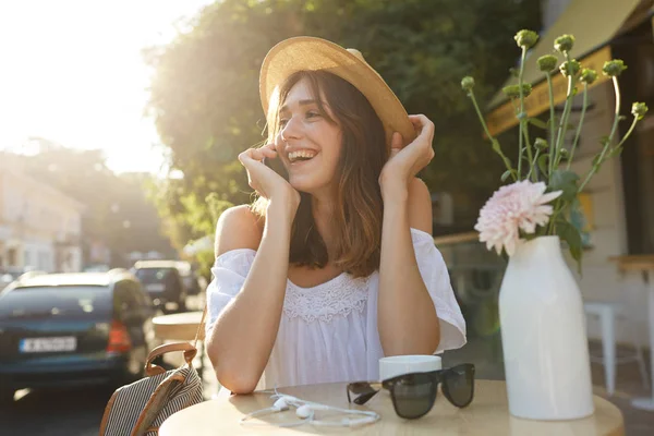 Cheerful attractive young woman wears summer hat and white blouse sitting in outdoor cafe, feels relaxed and talking on mobile phone in the city