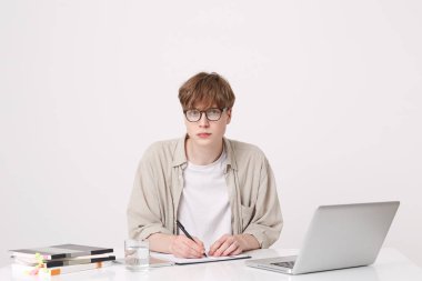 Ciddi yakışıklı genç öğrenci closeup bej gömlek dizüstü bilgisayar ve Notebook masa ve beyaz arka plan üzerinde izole yazma ile oturan gözlük takıyor