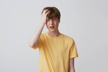 Portrait of desperate unhappy young man with hand on head and braces on teeth in yellow t shirt looks sad and having a headache isolated over white background clipart