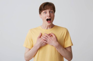 Açık ağız ve diş teli dişlerde şaşırmış sersem genç adam portresi sarı t gömlek tutar eller göğüs giyer ve beyaz arka plan üzerinde izole şaşırmış gibi görünüyor