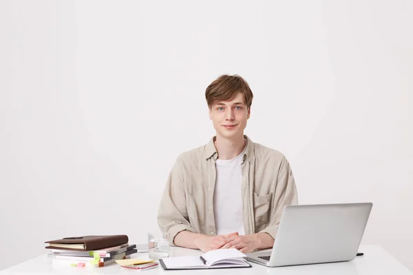 Portrait of handsome young man student wears beige shirt sitting at the table with laptop computer and notebooks isolated over white background