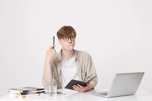 Portrait of pensive young man student wears beige shirt and glasses thinking and blowing bubbles with chewing gum at the table with laptop computer and notebooks isolated over white background