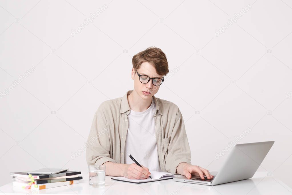 Portrait of serious young man student wears beige shirt and spectacles writing and study at the table using laptop computer and notebooks isolated over white background