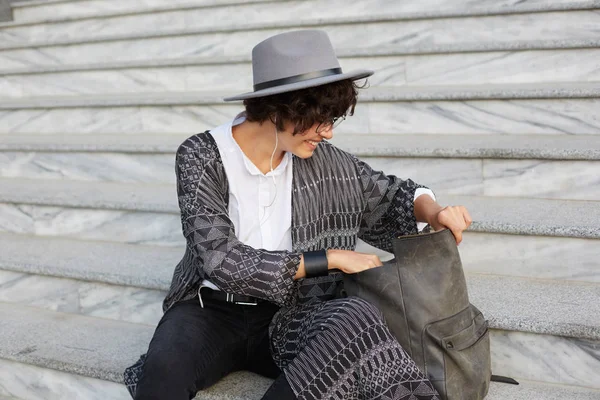 Young attractive female, wears cardigan, shirt, hat and glasses, with curly dark hair, open her backpack and searching something inside.