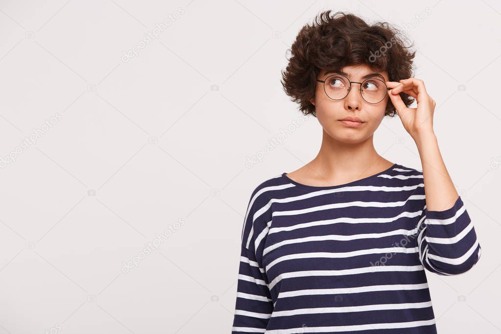 thoughtful young beautiful woman ponders the problem, looks aside, wears white and blue stripped sweatshirt and corrects or removes glasses, standing on the right side isolated over white background