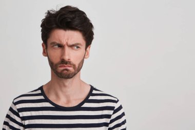 Young man with dark brown hair and beard wears black and white striped tshirt, looks angry, lips pursed frowning brows, head tuned rightside, stands leftside isolated over white background clipart