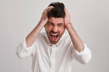 Sakallı genç brunet adam çığlık büyük bazı sorunlar nedeniyle, onun baş alın ve kaslar gerilir holding eller izole beyaz arka plan üzerinde beyaz klasik longsleave gömlek giyer görünüyor