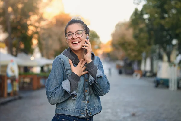 Beautiful lovely happy girl in glasses with blond hair gathered in the bun, smiles looking aside,wearing a denim jacket over sweatshirt, walking around the city talking with boyfriend on phone