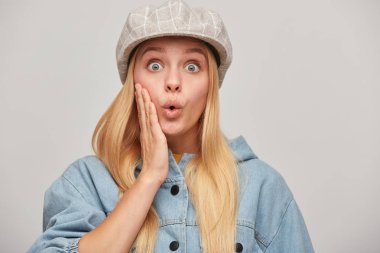 Emotional blonde young woman with hair down, looking very surprised pop-eyed, one hand holding her cheek, whistles in surprise, wears oversize denim jacket,beige checked cap, on grey background clipart