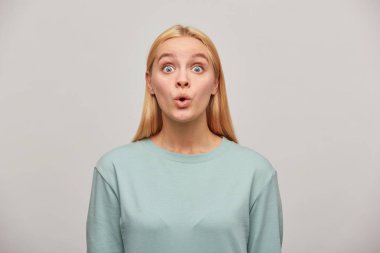 Emotional blonde young woman, looking very surprised, open-eyed, whistles in surprise, wearing blue casual sweatshirt, see something unexpected in front. Human reaction concept over grey background clipart