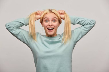 Uzun saç sürprizlerle, güzel genç sarışın kız kamerada, ekşitmeden vardır eğlenceli Şakalar, sevinir, gri arka plan üzerinde izole mavi rahat sweatshirt giyen başında, iki kuyruk yapar