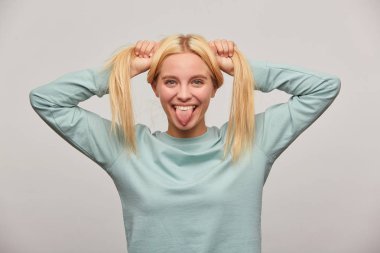 Dil, gösterilen serin komik süper kandırılan genç sarışın kız kamerada, ekşitmeden eğlenir, gri arka plan üzerinde izole mavi rahat sweatshirt giyen kafasında, iki kuyruk yapar