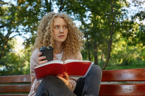 A young beautiful girl blonde sits with her legs tucked on a park bench, with coffee, notes, a notepad, looks away, thinks about something, trying to find a solution, bit her lower lip