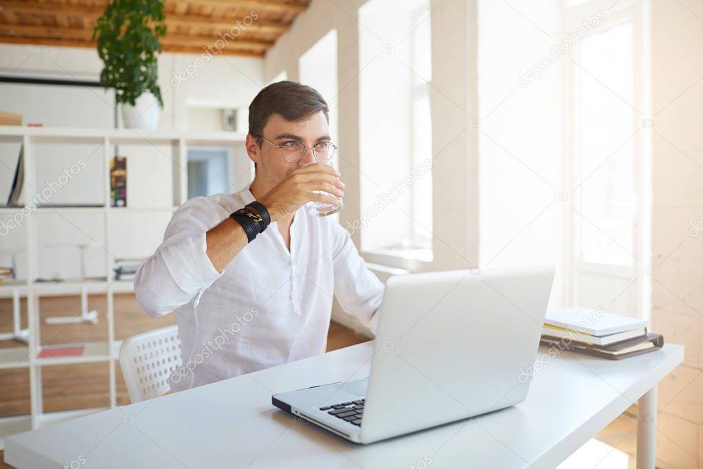 Closeup of attractive young businessman wears white shirt and spectacles working with laptop and drinking water at the table in office