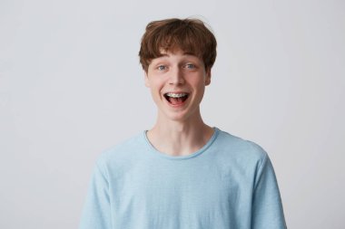 Close up of amazed excited young man with short disheveled hair and braces on teeth wears blue t-shirt shouting and feels happy surprised isolated over white background clipart