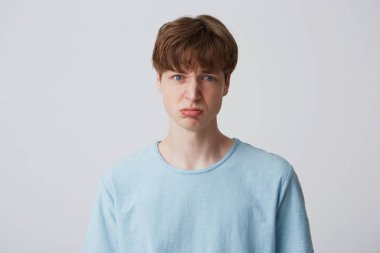 Portrait of blue-eyed young guy looks sad, upset, frustrated, displeased with the result of the test, lips pout, expresses insult, wears blue t-shirt stands isolated over white background. clipart