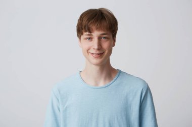 Close up of blue-eyed young guy with short haircut and braces on teeth wears blue t shirt slightly smiles and feels happy isolated over white background clipart