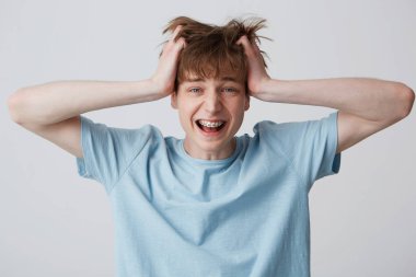 Screaming amazed excited young man clutched at his head, hair tousled, mouth wide opened as shouting loud with braces on teeth wears blue t-shirt, feels happy surprised isolated over white background clipart