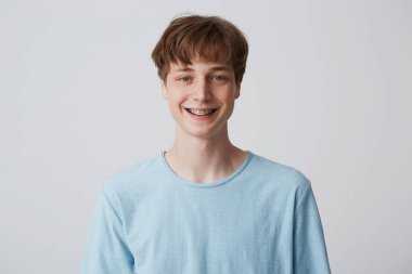 Teenager with short hair and braces on teeth looks camera, wears blue t-shirt, feels happy glad, smiling isolated over white background clipart