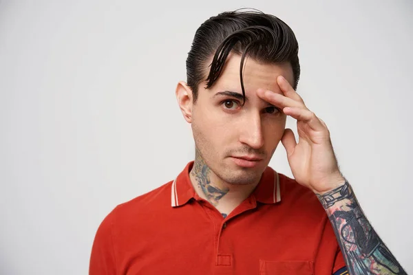 Portrait of a calm, self-confident guy, squinting at the camera, brown eyes, intriguing look, black hair, styling, one arm raised to the face, in a red polo t-shirt, has tattoos, on a white background
