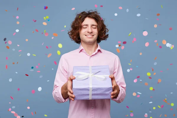 People, joy, fun and happiness concept. Relaxed happy birthday guy with eyes closed waiting for a surprise, posing for picture, holding box with present and with confetti falling down
