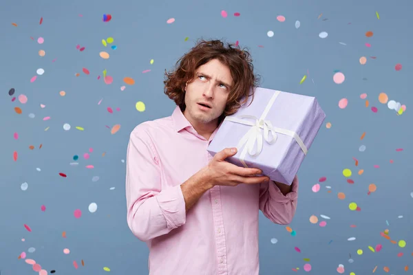 Close up of birthday man received a box as a gift, holds it in his hands near his head, looks away in the rumors about what\'s inside, confetti falling down. People, joy, fun and happiness concept.