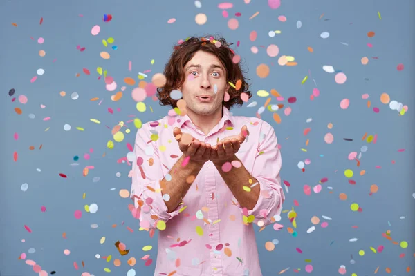 Young man blowing confetti off his hands. Studio shot of man happy standing around falling down confetti, over blue background