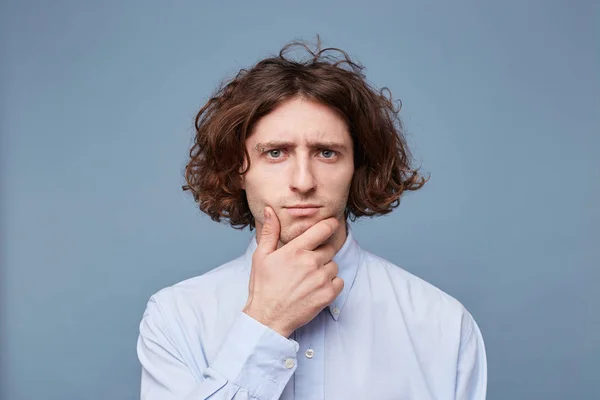 Portrait of a pensive young man dressed in shirt holding hand on his chin isolated over blue background. Creative and artistic male gazing thoughtfully at camera, thinking or making up ideas