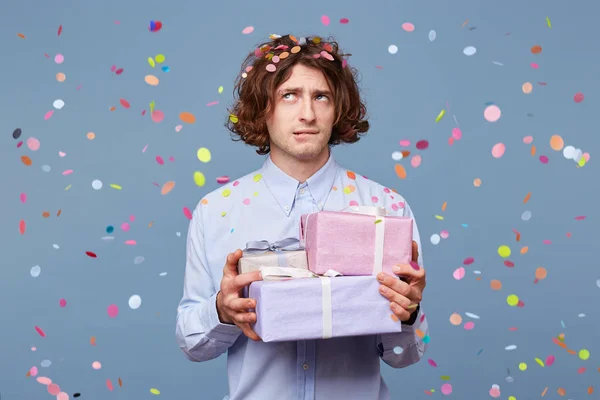 Young man accepts gifts for a birthday, holds boxes tied up with ribbons, looks thoughtfully puzzled upwards, bites lower lip, tries to remember the relatives\' names, confetti fall down