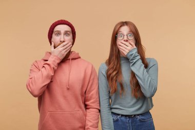 casualy, giyinmiş genç zencefil çift, görünüyor fotoğraf makinesi ile şok yüz ifadesi, büyük surprisement, içine kapalı atış ağız el ile kapatın. Biege'e arka plan üzerinde izole