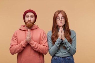 Genç çekici zencefil çift Biege'e arka planı, poz casualy giyinmiş ve jest dua ellerini tutun.