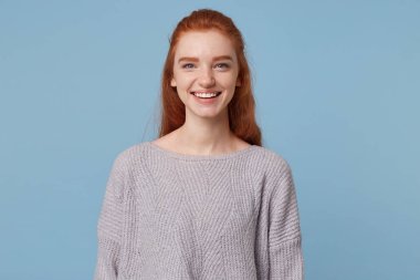 İhale güzel çekici bir kızıl saçlı kızı şirin portresi içtenlikle neşeyle affably gülümseyerek, dişlerini gösteren, mavi bir arka plan üzerinde izole bir büyük boy sıcak kazak giyiyor