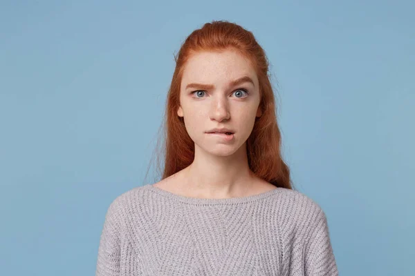 Young beautiful red-haired teen girl looks concerned puzzled in a panic, one eyebrow raised bit her lower lip isolated on a blue background.