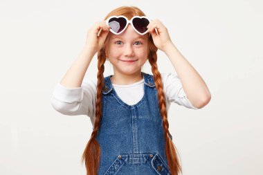 Neşeli mutlu neşeli Kızıl saçlı kız deniz üzerinde dinlenmek için gidiyor. Bir gülümseme ile kalp şeklinde giyer güneş gözlüğü. Beyaz bir arka plan üzerinde izole bir denim genel giymiş kameraya bakıyor.