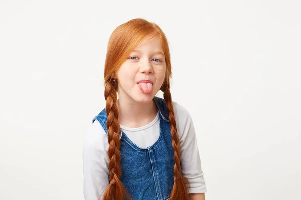 红头发的小女孩与辫子鬼脸在镜头显示舌头 女儿不想服从她的父母 女孩表现出她的侮辱 穿着牛仔裤工作服孤立在白色的背景 — 图库照片
