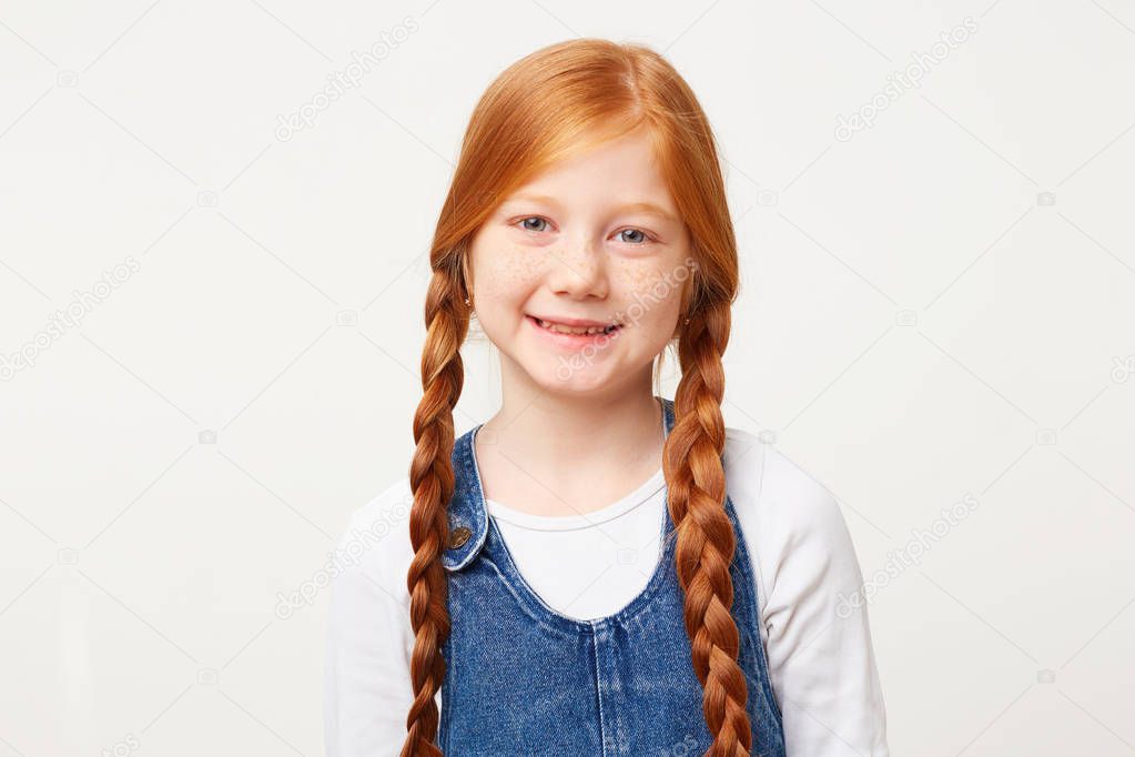 Portrait of sweet kind daughter with freckles and braided in two long plaits red hair, smiling nicely tenderly, dressed in jeans overall dress over a white longsleeve isolated on white background