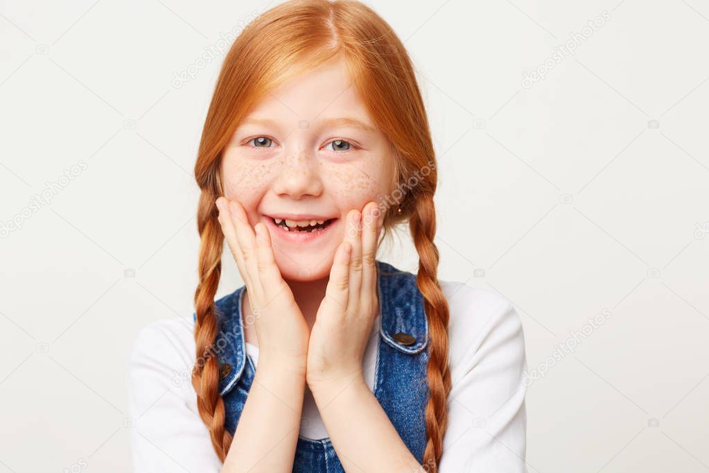 Close up portrait of smiling happy adorable girl with braided red hair in two long plaits, keeps palms near her face, dressed in jeans overall isolated on white background