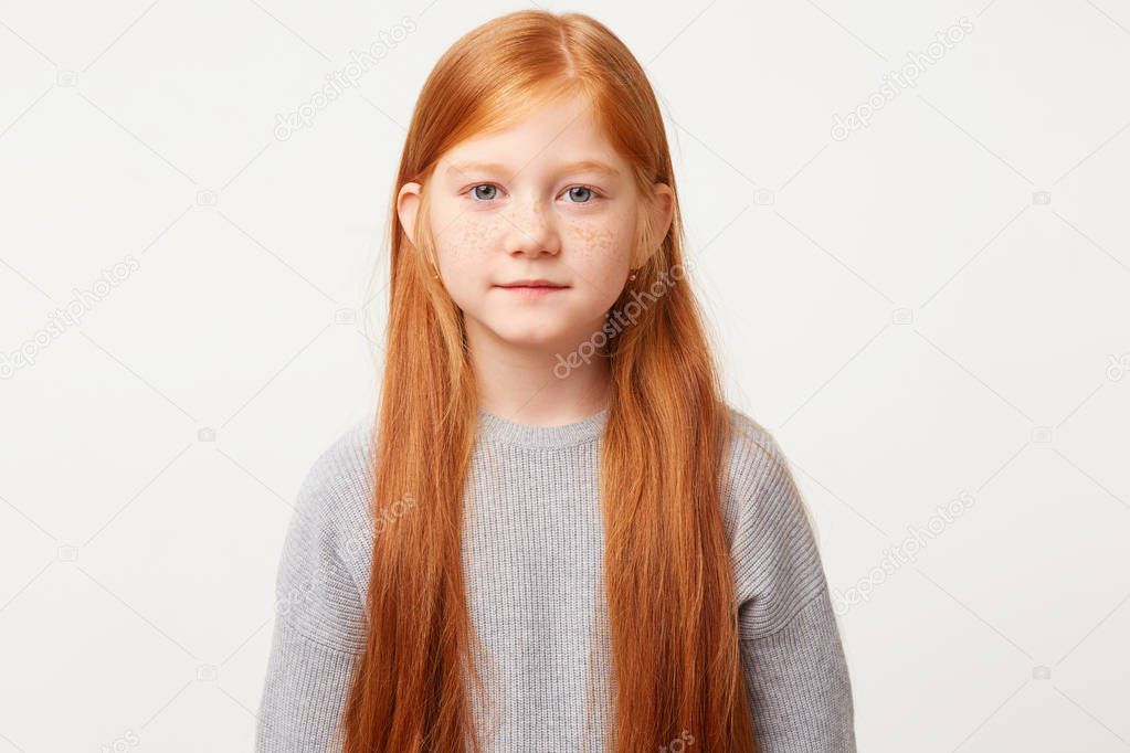 Cutie baby with long red hair down calmly looks into the camera. Beautiful girl behaves with restraint to observe the rules of behavior,over white background
