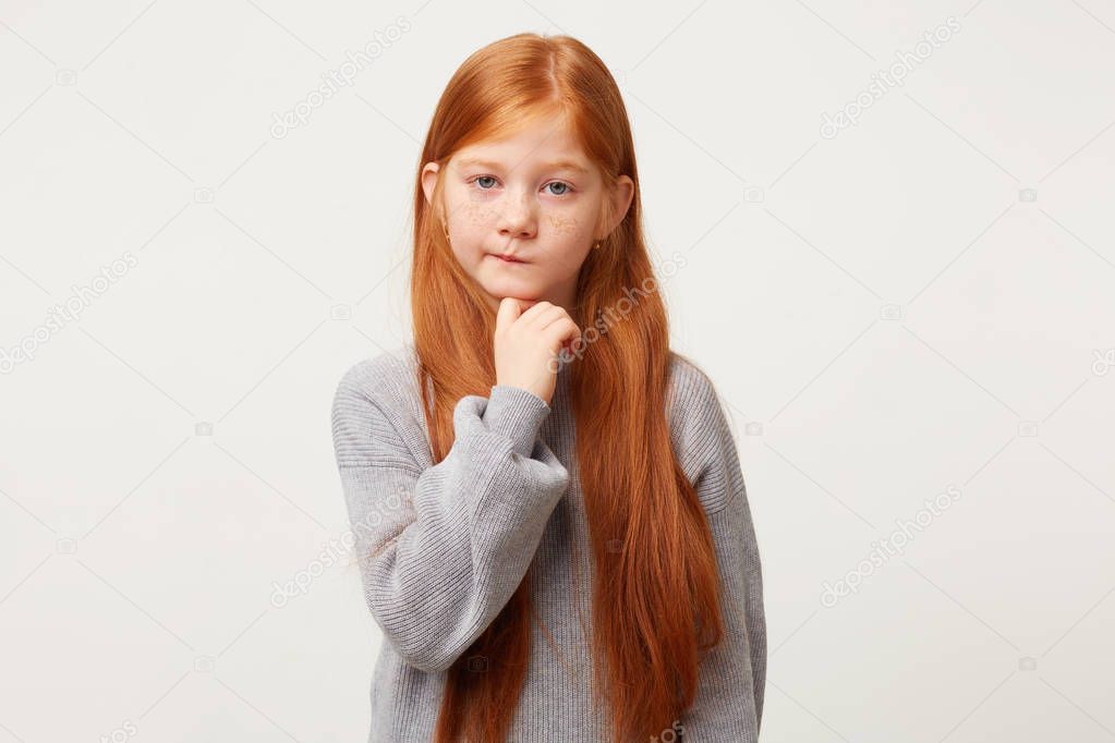 A little red-haired girl looks sadly upset, keeps fist near chin, lips disgruntledly pursed, looks thoughtful, thinks about something unreachable,dressed in gray sweater isolated on white background