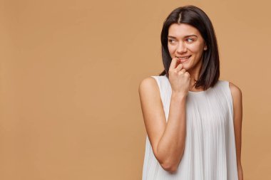 Mutlu heyecanlı düşünme bir şey hoş tabaklanmış kadın ayakta tutan parmağını ağzına sıcak gülümseme ile boş alanı sol tarafına bej renkli arka plan üzerinde görünüyor.
