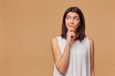 Unsatisfied thoughtful tanned woman standing hand holding chin, frowning, looking at the upper left corner at the blank copyspace, expressing suspiciousness and misunderstanding, over beige background clipart