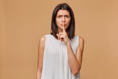 Endişeli, endişeli, endişeli, gergin, panik, agresif, öfkeli kadın, bej duvardan, sır olarak gizlilik için bir işaret parmağını ağzına aramalar tutan bir jest sessizlik, gösterir