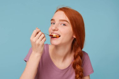 Güzel ve çekici Kızıl saçlı kız yiyor bir şey lezzetli tatma çalışır, kameraya görünüyor şakacı mavi bir arka plan üzerinde izole onun ağzına bir kaşık müsli ile koyar.