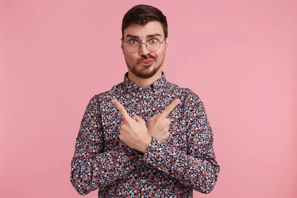 Young man in colorful shirt looking throught glasses having doubts and with confuse face expression keeps hands crossed ond pointing to bouth sides with inde fingers isolated on pink background — Stock Photo, Image