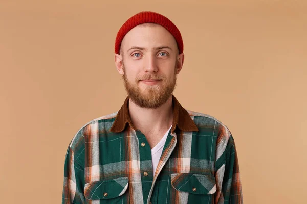 Un joven atractivo en el estudio mirando la cámara. Retrato de un tipo normal con amplia gorra roja y camisa a cuadros delante de fondo beige — Foto de Stock